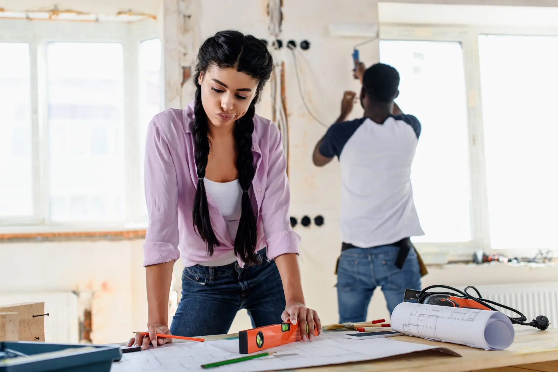 pensive young woman with spirit level looking at blueprint while her boyfriend working behind during